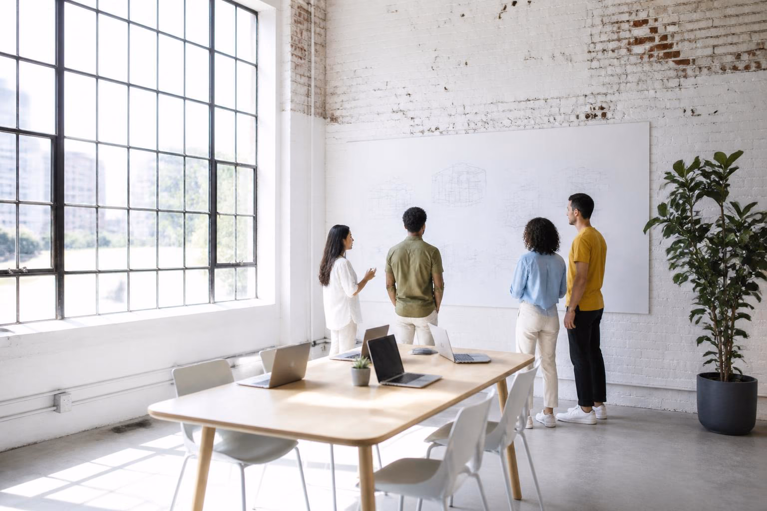 Four startup teammates standing at a whiteboard of architecture diagrams in a bright whitewashed Arts District brick loft with laptops on a long table