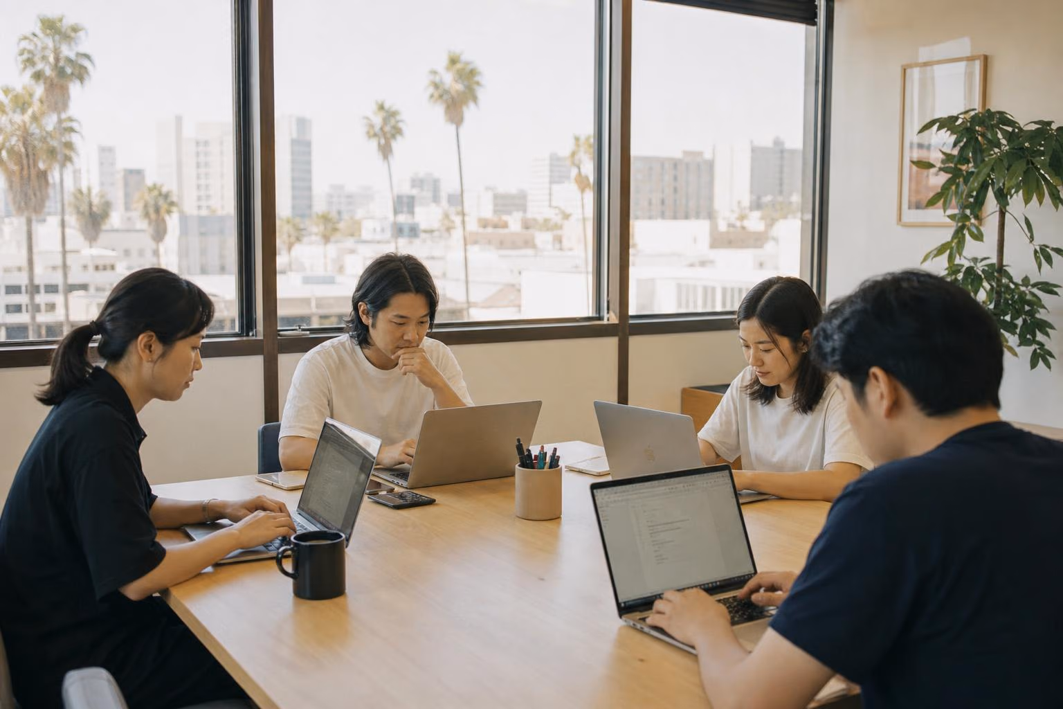 A small team working at laptops around a light wooden table in a bright Koreatown office, with natural daylight and palm trees visible outside.