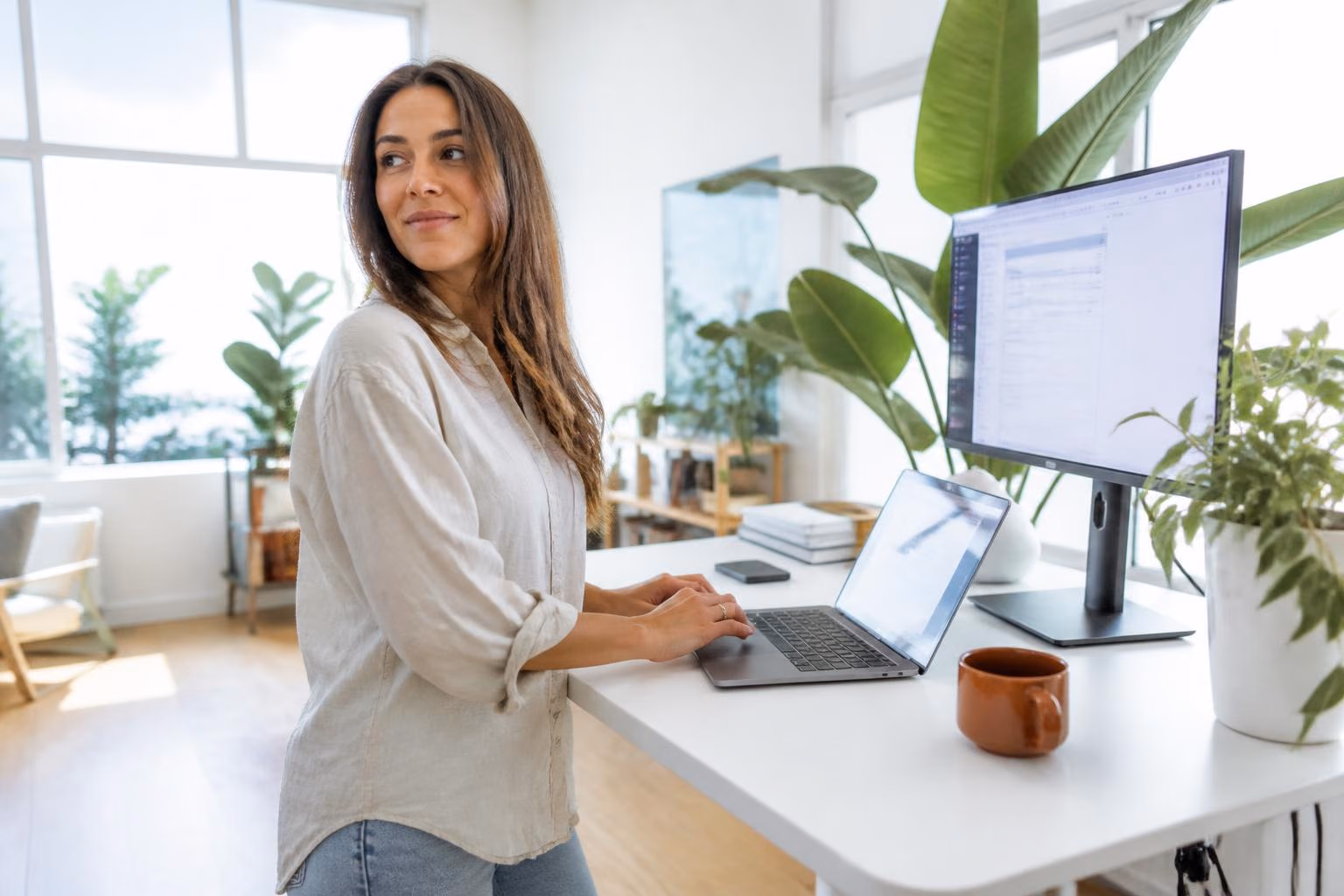 A woman in a linen shirt working at a white standing desk with a laptop and external monitor in a plant-filled, sunlit Culver City office